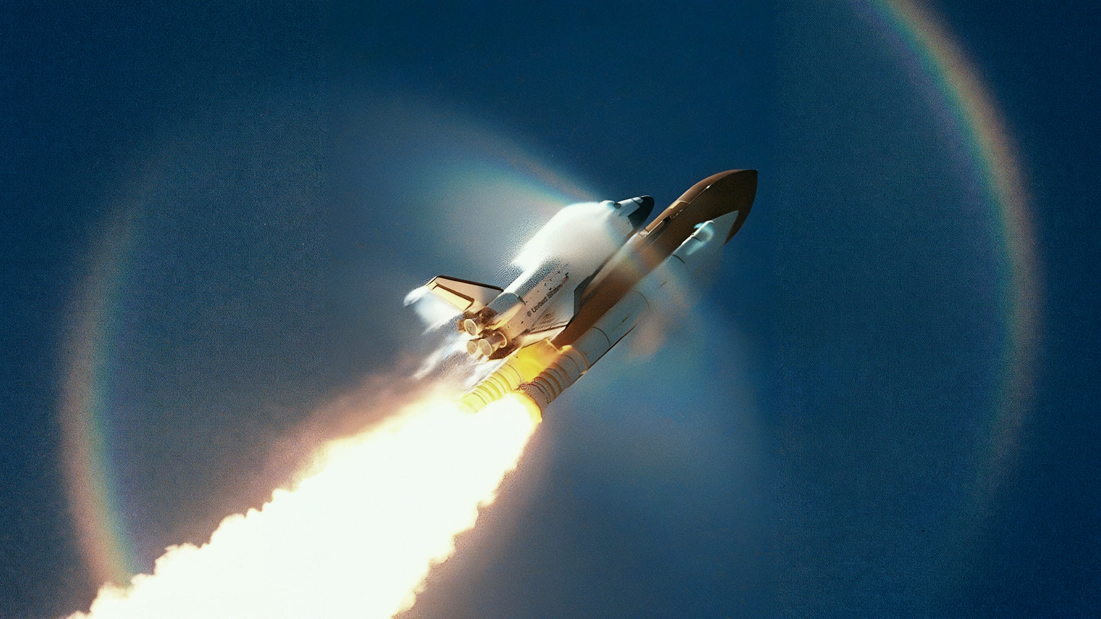 A space shuttle climbing through a deep blue sky on a trail of fire, circled by a faint rainbow halo.