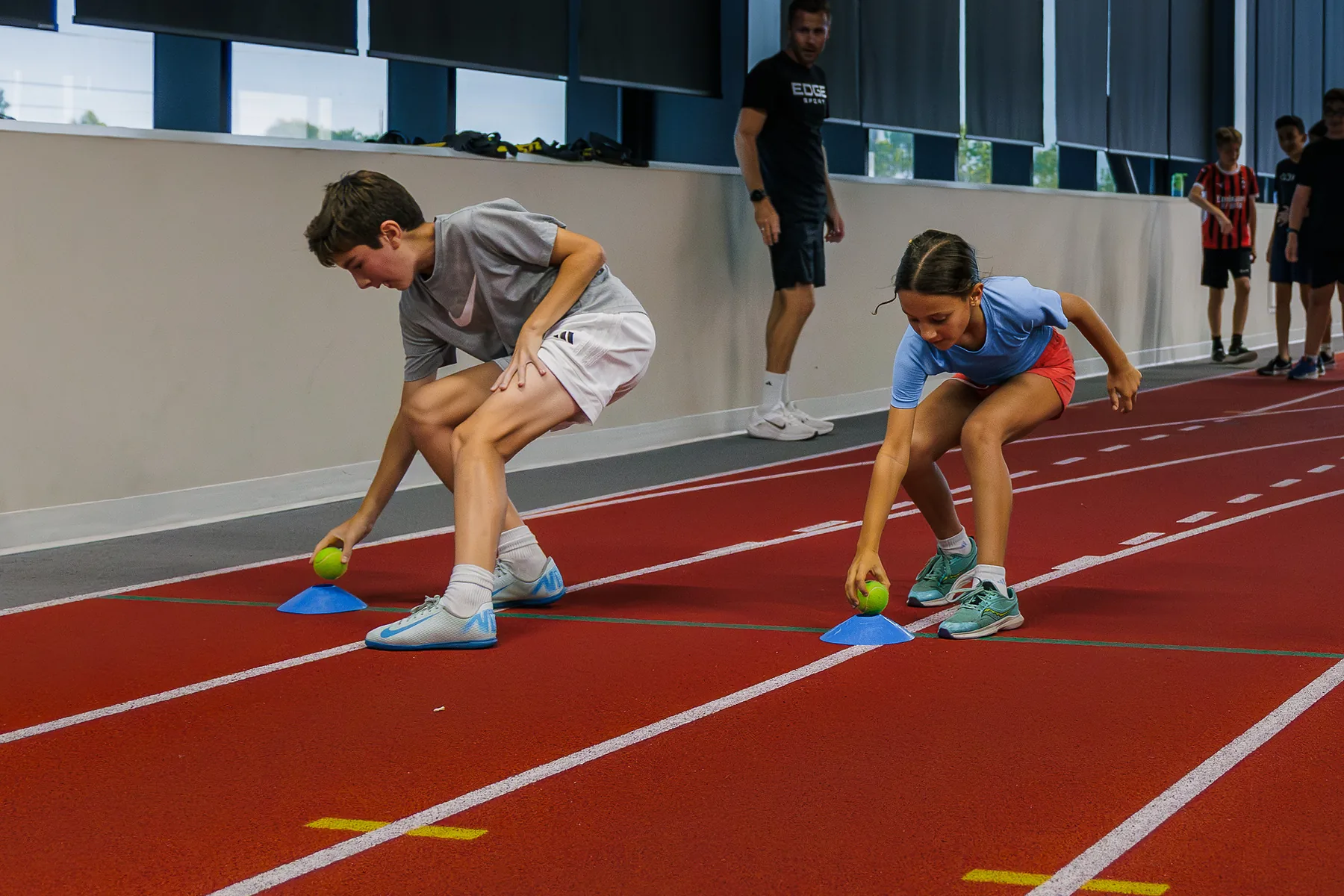Two young athletes picking up balls from cones on an indoor track