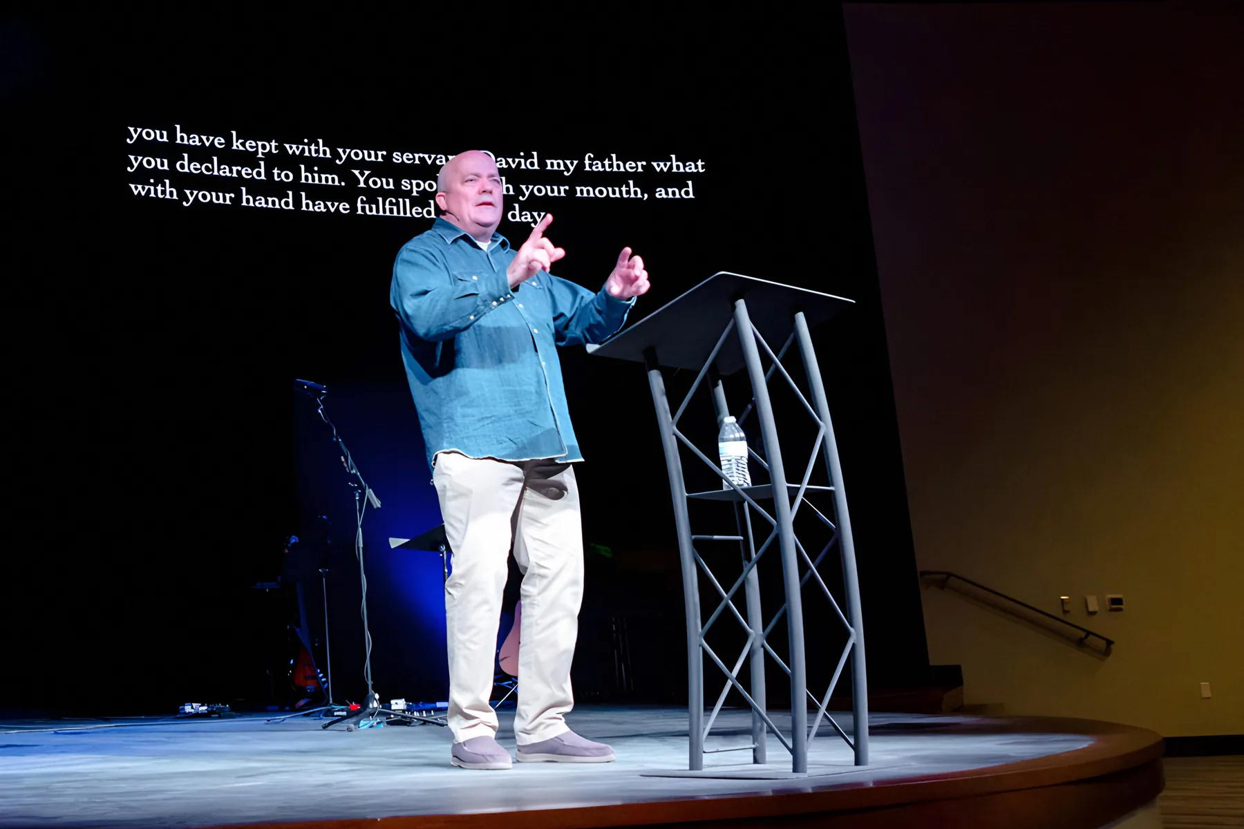 A pastor preaching on stage with scripture on the screen behind him