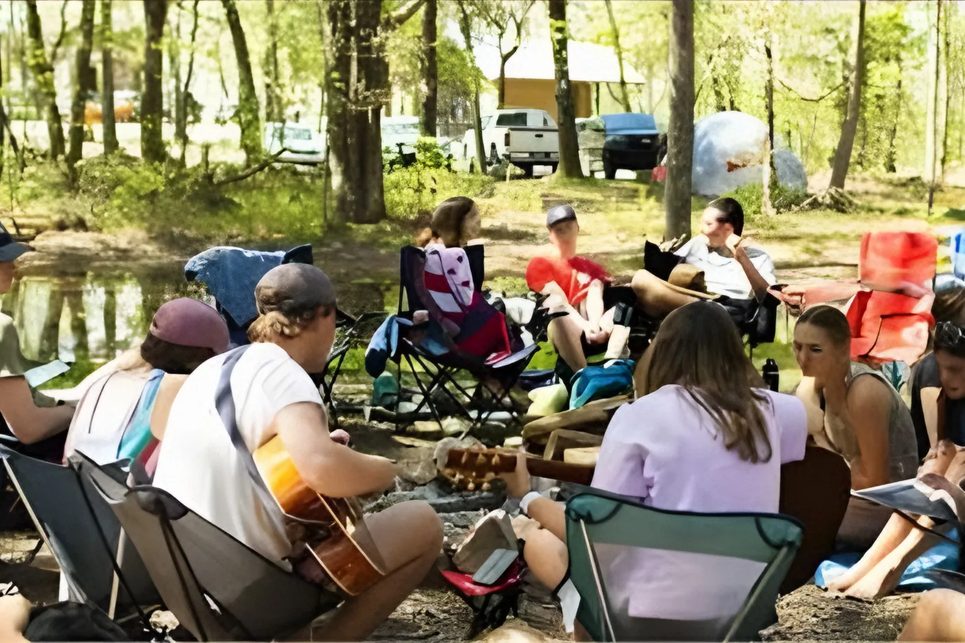 Club members in camp chairs by the water, one playing guitar